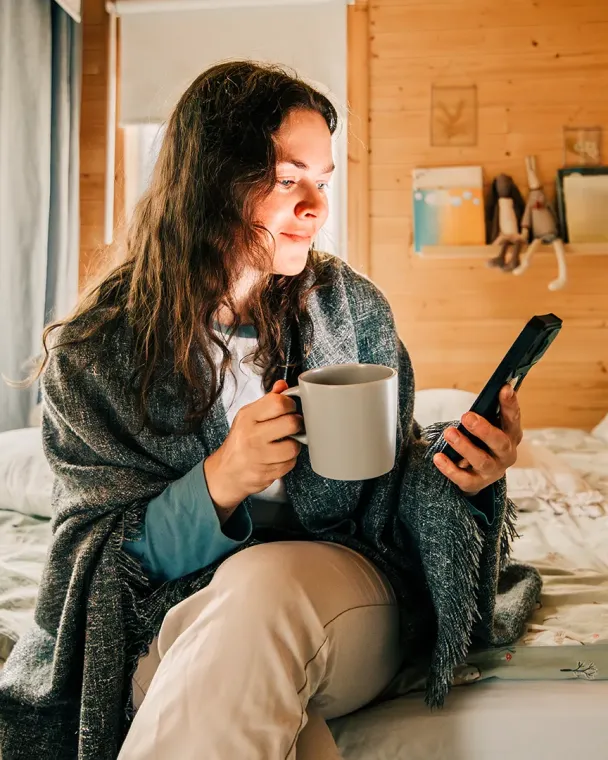 woman sitting on edge of bed with coffee in one hand and phone in the other